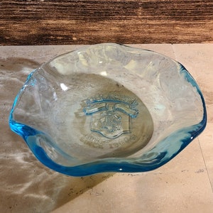 Op de afbeelding: Een lichtblauwe glazen schaal met een gegolfde rand. De schaal heeft een helder glazen embleem in het midden met de tekst "Old Orchard" en "Since 1904".