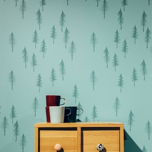 May include: A light oak cabinet with four drawers, each with a different decorative knob. The cabinet is set against a teal wall with a repeating pattern of stylized evergreen trees. Three mugs in red, white, and dark teal are stacked on top.