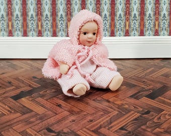 Può includere: Una bambola in miniatura con carnagione chiara e occhi scuri, che indossa una mantella con cappuccio in maglia rosa chiaro con chiusura a laccio e pantaloni in maglia rosa chiaro abbinati. La bambola è seduta in posizione frontale e morbida.