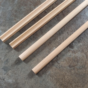 Könnte beinhalten: Vier unfertige Holzleisten in verschiedenen Formen und Größen sind diagonal auf einer melierten grauen Oberfläche angeordnet. Zwei Leisten haben ein abgerundetes Profil, während die anderen beiden ein gerilltes Design aufweisen. Das Holz hat eine helle, natürliche Farbe.