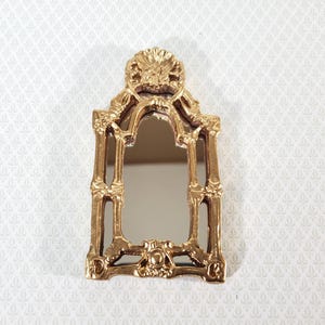 May include: A miniature gold-colored mirror with an ornate, baroque-style frame. The frame features intricate detailing, including floral and scrollwork designs. The mirror has a rectangular shape with an arched top, suitable for dollhouses or decorative displays.
