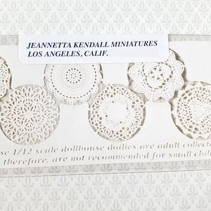 May include: A package of white, 1/12 scale dollhouse doilies in various lace patterns. The package includes a label that reads "JEANNETTA KENDALL MINIATURES LOS ANGELES, CALIF." and a warning about small children.