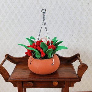 May include: A miniature terracotta-colored hanging planter filled with red and white flowers and green leaves. The planter is suspended by a silver wire hanger and sits on a small, dark wooden table. The table has a drawer and a decorative handle.