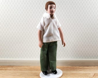 Può includere: Una bambola in miniatura con capelli castani corti e carnagione chiara, che indossa una camicia bianca a maniche corte con colletto e pantaloni verde scuro con una sottile trama diagonale. La bambola è su una base circolare bianca e indossa scarpe nere.