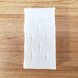 May include: A rectangular white cushion with a tufted design. The cushion is made of a soft, textured fabric and has a series of evenly spaced, recessed squares. The cushion is placed on a wooden surface.