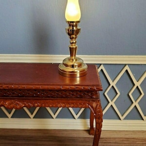 May include: A miniature brass oil lamp with a frosted glass shade is lit, casting a warm glow. The lamp sits on a small, intricately carved wooden table with claw feet. The table is a rich, dark brown color.