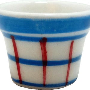 May include: A small, white ceramic cup with a blue band around the top and horizontal blue stripes. Vertical red lines are painted on the cup. The cup has a slightly flared shape.