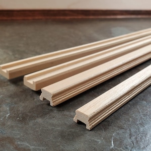 May include: Close-up of several unfinished wooden molding pieces. The molding has a decorative profile with a flat top and a series of grooves along the side. The wood has a light, natural color and is ready for staining or painting.