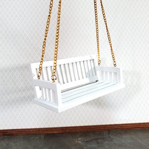May include: A miniature white wooden swing with a slatted back and seat, suspended by gold-colored chains. The swing is designed for decorative purposes and is set against a patterned wall.