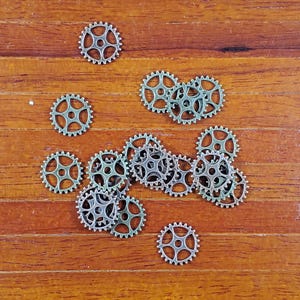 May include: A collection of small, metallic gears scattered on a wooden surface. The gears have a circular shape with multiple teeth around the edges and a central hole. The gears are a silver color.