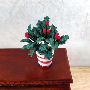 May include: A miniature potted plant with green leaves and red berries. The plant is in a white pot with red stripes. The pot is sitting on a dark brown wooden surface.