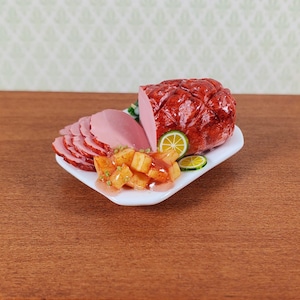 May include: A miniature food display on a white rectangular plate featuring a glossy, dark red glazed ham, several thin pink slices of ham, a pile of diced orange fruit or vegetables, and two thin, round slices of green lime as a garnish.