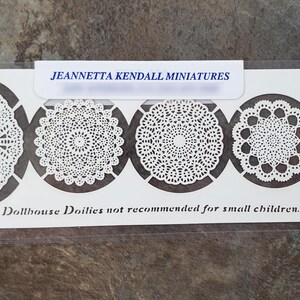 May include: A package of miniature dollhouse doilies. The white doilies feature intricate lace designs in oval and circular shapes. The text "JEANNETTA KENDALL MINIATURES" is at the top. The text "Dollhouse Doilies not recommended for small children" is at the bottom.