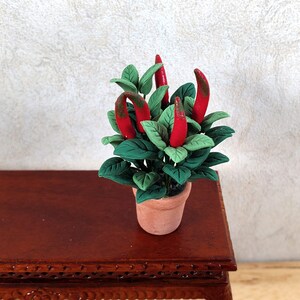 May include: A miniature clay plant with bright red chilli peppers and green leaves in a small terracotta pot. The plant sits on a dark brown wooden table, with a textured white wall in the background.