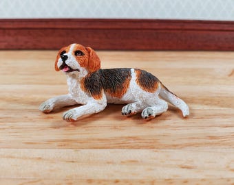 May include: A miniature beagle figurine, lying down with its tongue slightly out. The dog has a white, brown, and black coat, with detailed paws and a realistic expression. The figurine is set on a wooden surface.