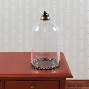 May include: A clear glass cloche with a decorative metal base and top. The cloche sits on a wooden surface. The base has a scalloped edge. The top has a small, round, brass-colored handle.
