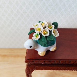 May include: A miniature ceramic turtle planter with a turquoise rim, filled with white flowers and green leaves. The turtle is white with yellow feet and a black eye. The planter is on a dark brown wooden surface.