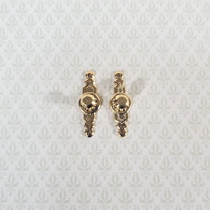 May include: Two identical gold-toned metal drawer pulls featuring an elongated, symmetrical shape with a central circular knob and decorative geometric engravings along the vertical body, designed for attachment with a single screw hole in the center.