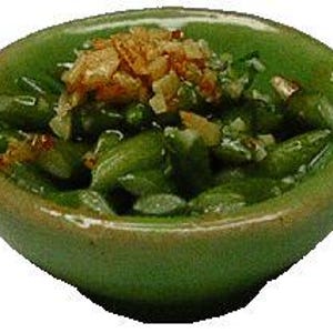 May include: A small, round, green ceramic bowl filled with green beans and topped with a garnish. The bowl has a slightly uneven rim and a darker green trim. The garnish appears to be fried onions or a similar crispy topping.