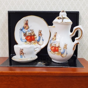 May include: A miniature white porcelain tea set featuring Peter Rabbit illustrations with gold trim. The set includes a teapot with a lid, a teacup, and two saucers, all decorated with colorful rabbit characters. The items are displayed in a black box.