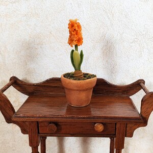 May include: A miniature orange hyacinth flower in a terracotta pot sits on a small, ornate wooden table. The table has a drawer and decorative handles. The background is a textured, off-white wall.