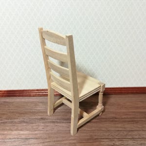 May include: A miniature wooden chair with a light, natural finish. The chair has a slatted back and a simple, rectangular seat. The legs are straight with a decorative turned detail. The chair is designed for a dollhouse or miniature setting.
