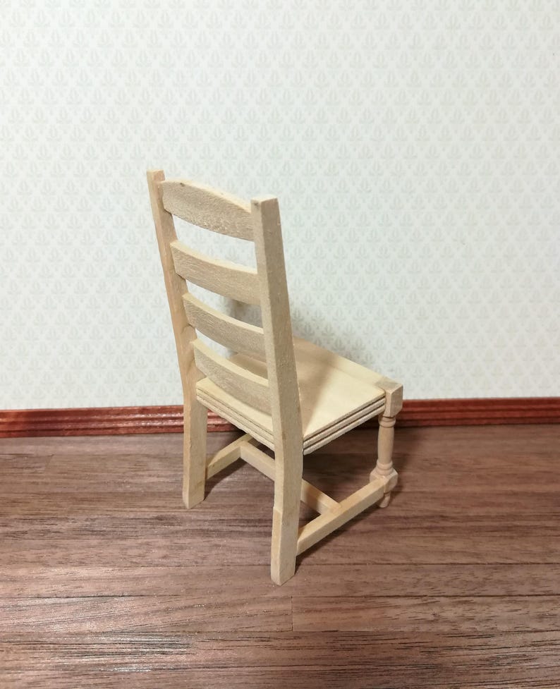 May include: A miniature wooden chair with a light, natural finish. The chair has a slatted back and a simple, rectangular seat. The legs are straight with a decorative turned detail. The chair is designed for a dollhouse or miniature setting.