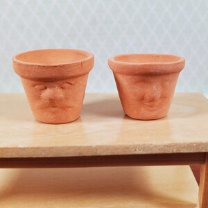 May include: Two small, terracotta-coloured flower pots with a face design. The pots are sitting on a light-coloured wooden table. The pots have a round opening and a slightly textured surface.