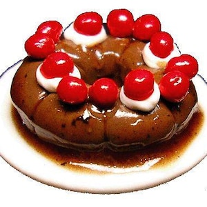 May include: A miniature chocolate cake with a ring shape, topped with bright red cherries and white cream, sits on a white plate. The cake is glazed with a glossy, brown sauce. A dessert item.