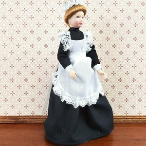 May include: A miniature doll dressed as a maid, wearing a black dress, white apron, and a lace-trimmed cap. The doll has a painted face with red lips and brown hair. The doll is standing on a wooden surface.