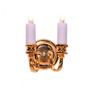 May include: A miniature gold-colored wall sconce with two white candle-shaped lights. The sconce has a round base and curved arms, with intricate detailing. The lights are illuminated, creating a warm glow.