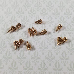 May include: A collection of small, gold-colored metal decorative beads. Each bead has a unique shape with a rounded top and a pointed bottom, and a small post. The beads are scattered on a patterned, light-colored surface.