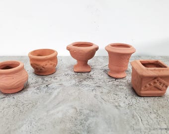 Könnte beinhalten: Eine Sammlung von fünf kleinen Terrakottatöpfen in verschiedenen Formen, darunter abgerundete, kelchförmige und quadratische Designs. Jedes Miniaturgefäß hat eine natürliche rotbraune Tonoberfläche mit dezenten Prägemustern an den Außenseiten.
