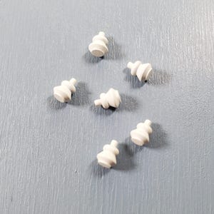 May include: A collection of six small, off-white, matte-finished ceramic or plastic components, each featuring a tiered, rounded shape with a narrow cylindrical stem at the top, arranged in a scattered pattern on a textured grey surface.