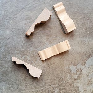 May include: Four unfinished wooden decorative molding pieces. Three pieces have a curved, scalloped edge, and one is a rectangular block. The pieces are light-colored and lie on a mottled gray and brown surface.
