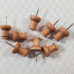 May include: A collection of seven wooden push pins with silver metal tips. The push pins are a light brown color and are arranged on a patterned, light gray background. The push pins are a stationery item.