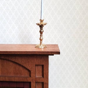 Könnte beinhalten: Ein goldfarbener Kerzenhalter mit einer weißen Kerze steht auf einem hölzernen Kaminsims. Der Kerzenhalter hat ein verziertes Design und einen quadratischen Sockel. Der Kaminsims ist aus dunklem Holz gefertigt und hat eine dekorative Platte. Der Hintergrund ist eine gemusterte Tapete.