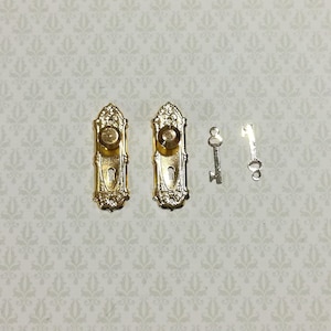 May include: Two miniature gold-colored metal door plates with round knobs and keyholes, accompanied by two small silver-colored metal keys. The door plates feature an ornate, embossed design with a vertical rectangular shape and rounded ends.