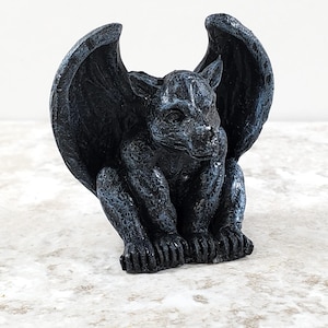 May include: A detailed, black gargoyle statue with large, textured wings. The mythical creature is seated, with its wings spread wide. The statue has a weathered appearance, with hints of gray highlighting the details. A decorative item for gothic or fantasy themes.