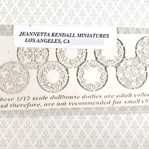 May include: A package of miniature dollhouse doilies. The white paper doilies are in various circular designs. The package includes text that reads "JEANNETTA KENDALL MINIATURES LOS ANGELES, CA". The doilies are 1/12 scale and are adult collectibles.