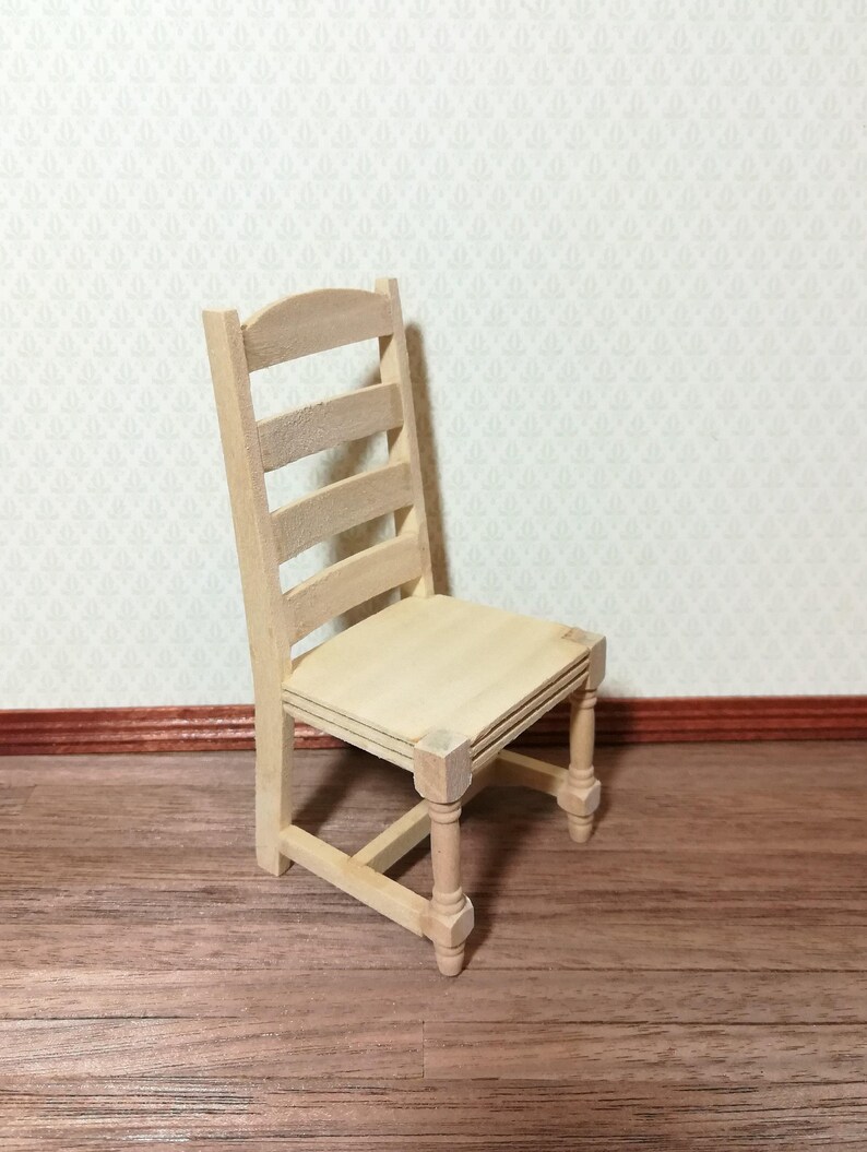 May include: A miniature wooden chair with a ladder back and a square seat. The chair is unfinished, showing the natural wood grain. The chair's dimensions are approximately 6.35 cm tall.
