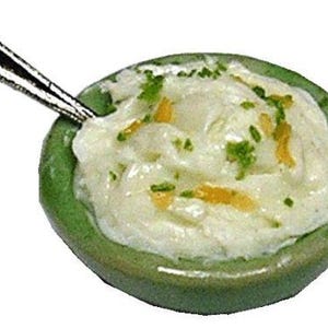 May include: A miniature green ceramic bowl filled with a creamy white dip, garnished with green and yellow flecks. A small silver spoon rests in the dip, suggesting a serving of a savory appetizer.