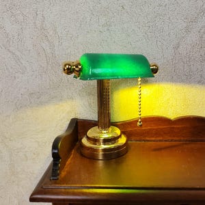 May include: Miniature green glass and gold-toned desk lamp. The lamp has a classic banker's lamp design with a green glass shade and a gold-colored base and stem. The lamp is sitting on a wooden surface.
