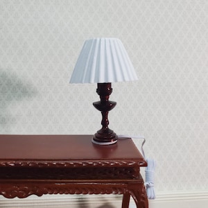 May include: Miniature table lamp with a white pleated lampshade and a dark brown wooden base. The lamp is placed on a dark brown wooden table. The lamp has a classic design and is suitable for a dollhouse or miniature setting.
