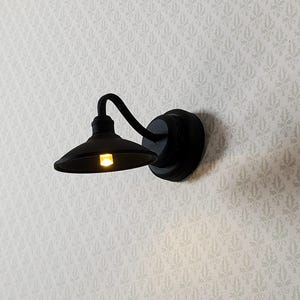 May include: A miniature black wall-mounted light fixture with a curved arm and a small, round shade. The light is on, emitting a warm, yellow glow. The fixture is mounted on a wall with a subtle, repeating pattern.