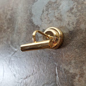 May include: A gold-colored tie clip with a rounded base and a rectangular bar. The clip has a simple, elegant design, perfect for securing a tie. The metal has a shiny finish.