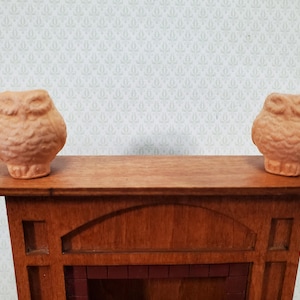 May include: Two small, light brown, textured owl figurines made of a matte material, each positioned on one side of the top surface of a miniature wooden fireplace mantel that features a dark red brick-like interior detail.