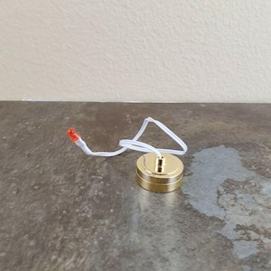 May include: A small, gold-coloured, round electrical component with a white wire and a red light at the end. The component is on a grey surface, with a white wall in the background. The item appears to be a small light fixture component.