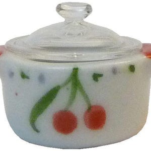 May include: A small, white ceramic casserole dish with a clear glass lid. The dish features a hand-painted cherry design with red cherries and green leaves. The handles are tipped with red paint.