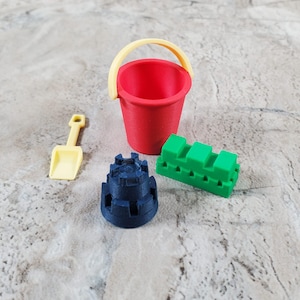 May include: A red bucket with a yellow handle, a yellow shovel, a dark blue castle, and a green block. These colorful toys are designed for sand play and are suitable for children.